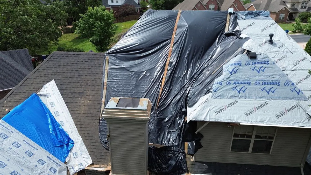 Black tarp and synthetic underlayment covering a damaged shingle roof beside a chimney