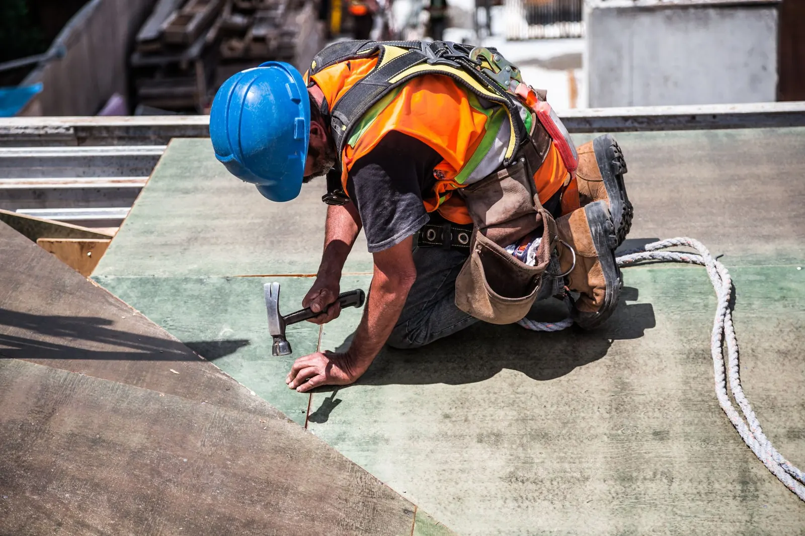 Roofer in a hard hat and safety harness nailing down a low-slope roof deck