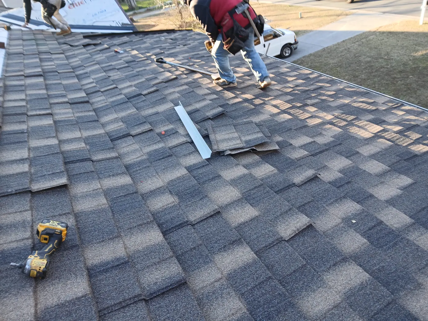 A roofer laying architectural shingles course by course, with a cordless drill and a strip of metal flashing on the roof