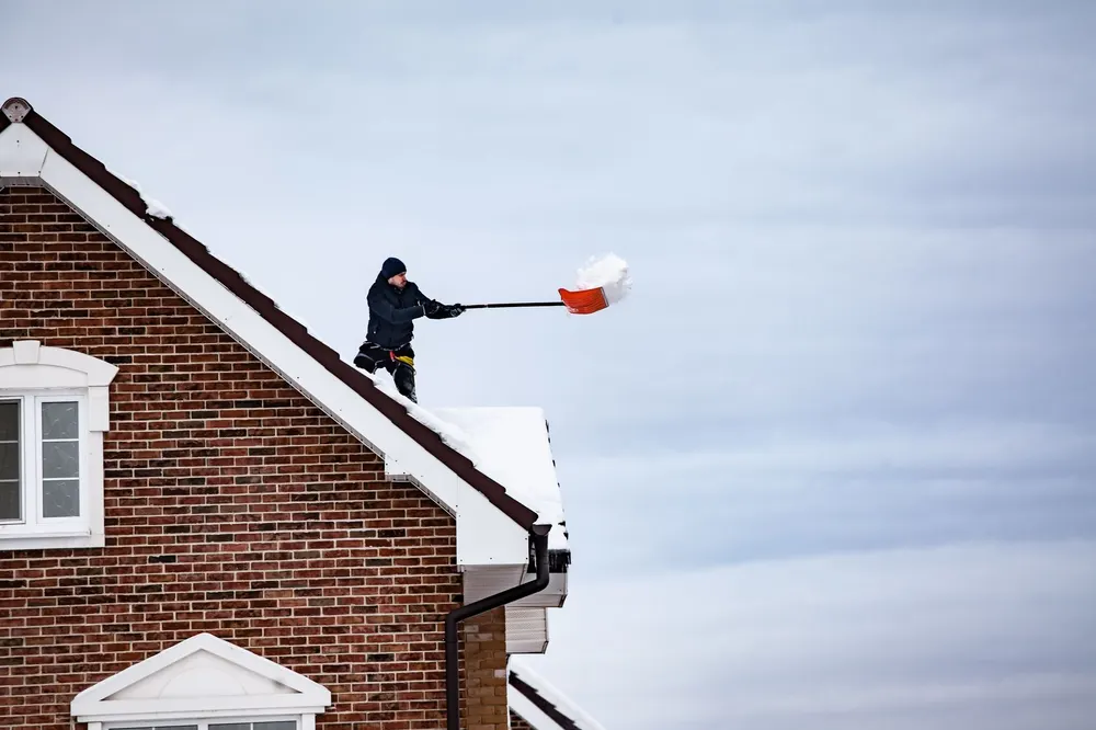 A man standing on the snowy roof of a brick house, throwing snow off with a red shovel