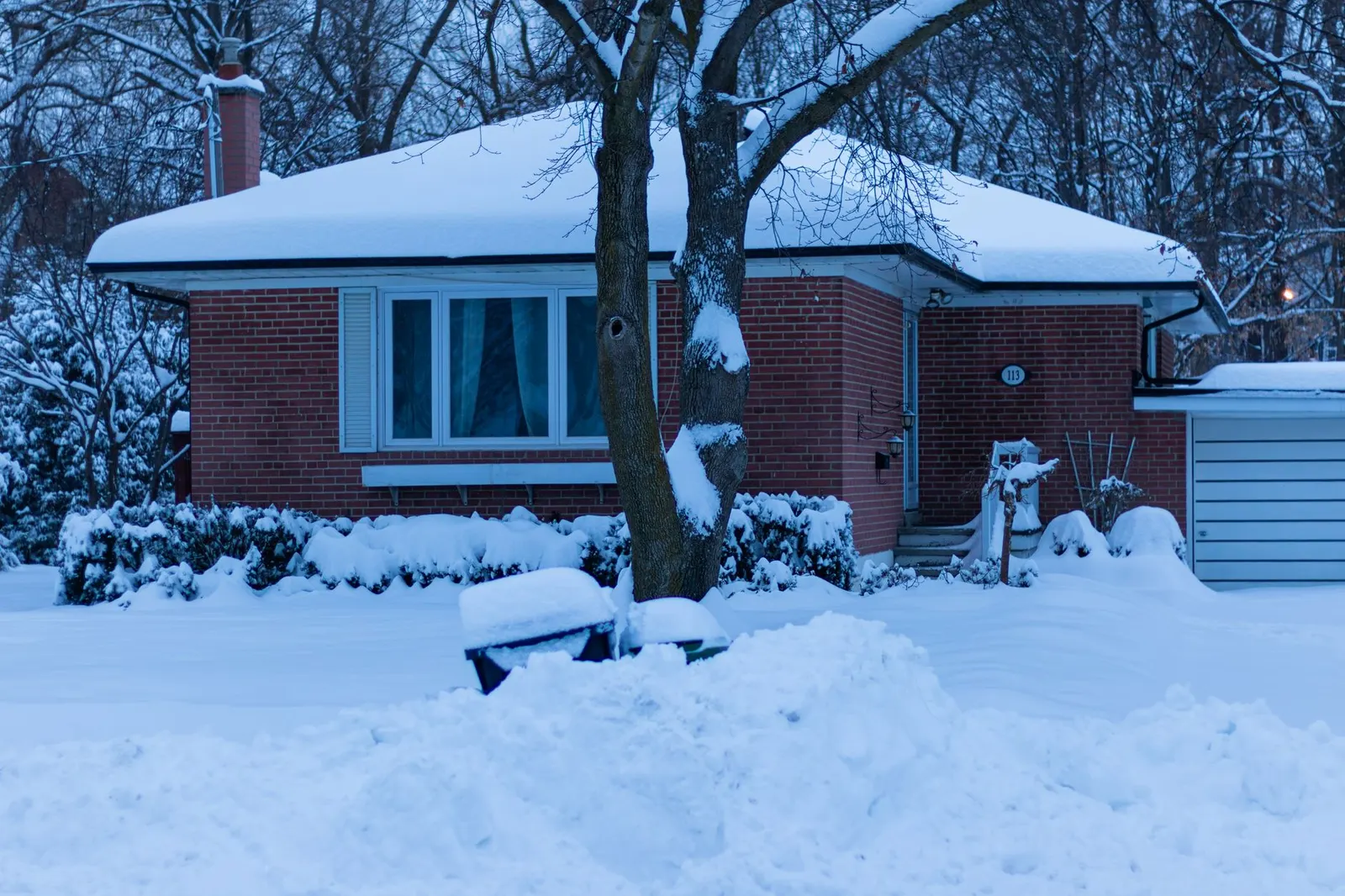Brick bungalow with deep snow on its hip shingle roof and in the yard, bare trees behind it