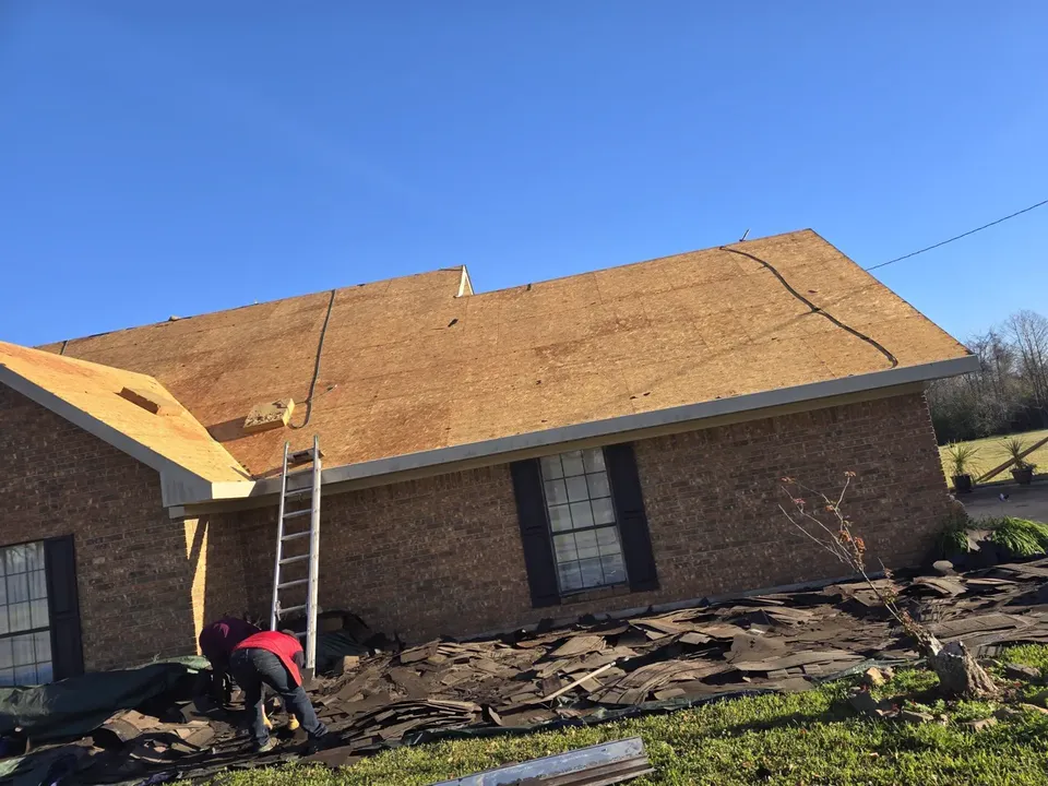 Brick house with its old shingles stripped to bare decking, a ladder at the eave and torn-off shingles on tarps below